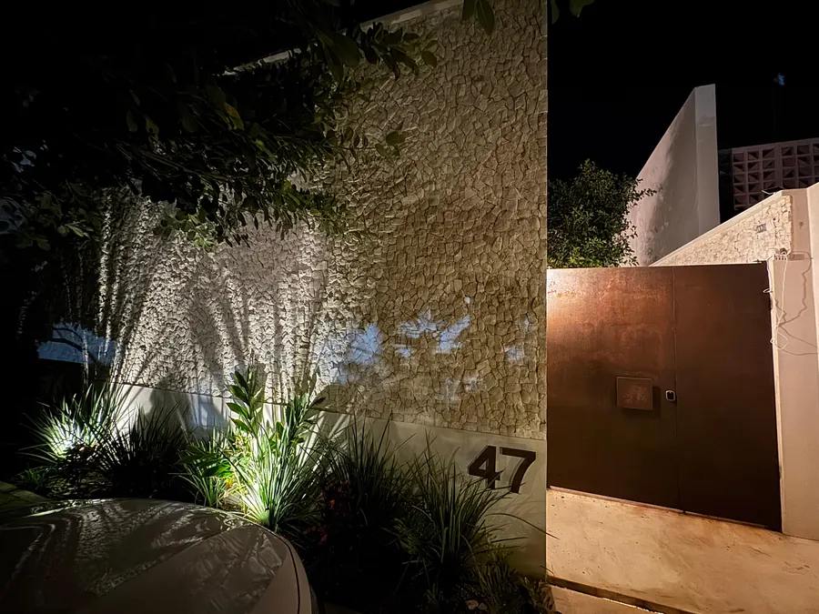 Fachada de noche de la casa en Provincia Golf Club: muro de chukum iluminado y portón de acero cortén