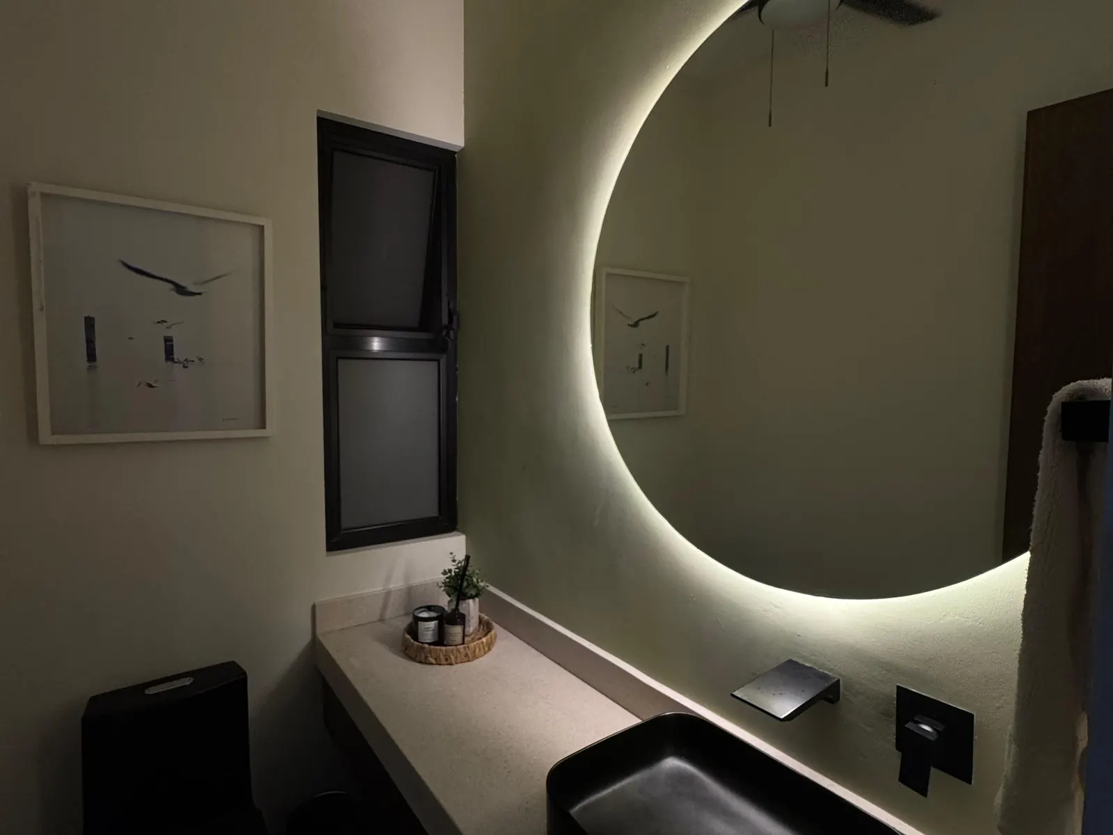 Baño con espejo circular retroiluminado, lavabo negro y cubierta de concreto