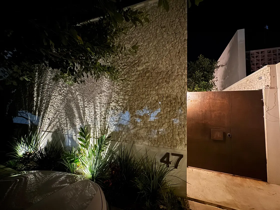Fachada de noche: muro de chukum iluminado desde el piso, número 47 y portón de acero cortén